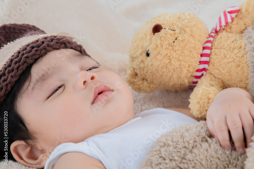Asian adorable newborn baby wear brown knit hat deeply sleeping with beige blanket next to teddy bear and toy camera with safe and comfortable. Little toddler cute resting after eat full.