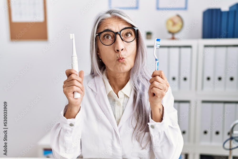 Middle age grey-haired woman working at dentist clinic holding electric teethbrush and toothbrush smiling looking to the side and staring away thinking. Middle age grey-haired woman working at dentist clinic holding electric teethbrush and toothbrush smiling looking to the side and staring away thinking.