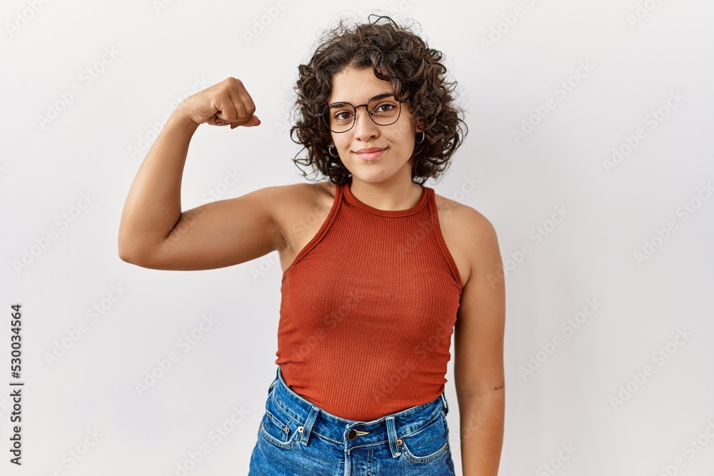 Young hispanic woman wearing glasses standing over isolated background strong person showing arm muscle, confident and proud of power Young hispanic woman wearing glasses standing over isolated background strong person showing arm muscle, confident and proud of power