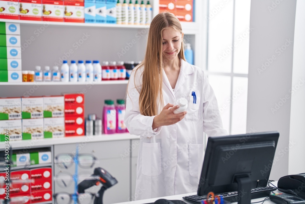 Young caucasian woman pharmacist holding pills bottle using computer at pharmacy