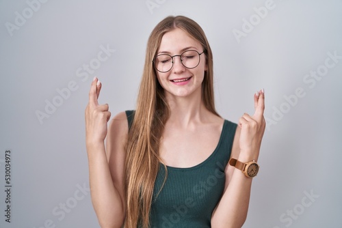 Young caucasian woman standing over white background gesturing finger crossed smiling with hope and eyes closed. luck and superstitious concept.
