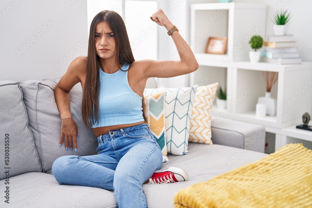 Young brunette woman sitting on the sofa at home strong person showing arm muscle, confident and proud of power Young brunette woman sitting on the sofa at home strong person showing arm muscle, confident and proud of power