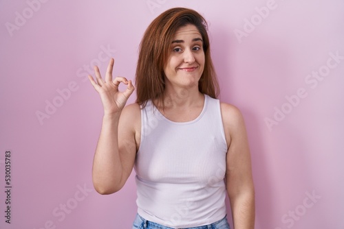 Brunette woman standing over pink background smiling positive doing ok sign with hand and fingers. successful expression.