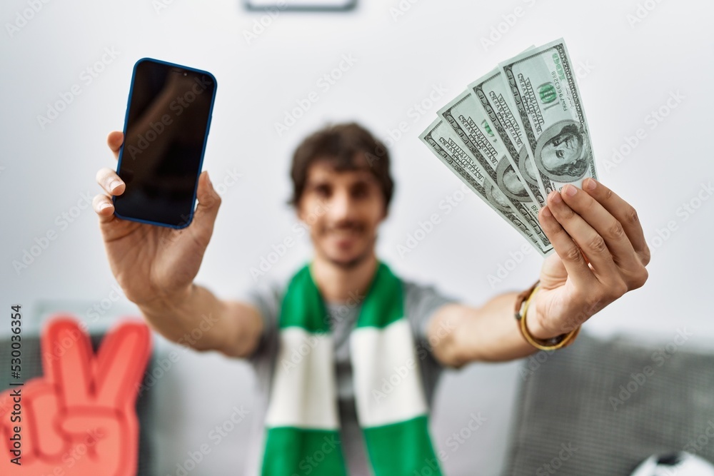 Naklejka premium Young hispanic man holding smartphone and dollars at home
