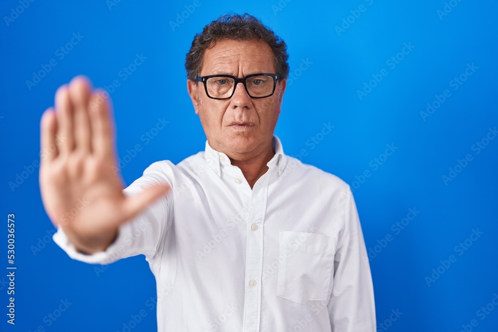 Middle age hispanic man standing over blue background doing stop sing with palm of the hand. warning expression with negative and serious gesture on the face. Middle age hispanic man standing over blue background doing stop sing with palm of the hand. warning expression with negative and serious gesture on the face.