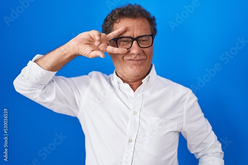 Middle age hispanic man standing over blue background doing peace symbol with fingers over face, smiling cheerful showing victory