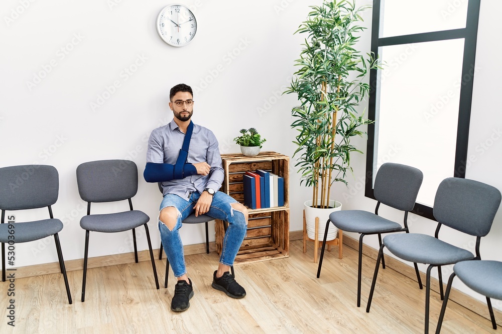 Young arab man sitting on chair with arm sling at clinic waiting room Young arab man sitting on chair with arm sling at clinic waiting room