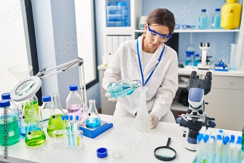 Young hispanic woman wearing scientist uniform measuring liquid at laboratory