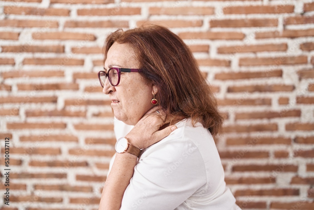 Senior woman with glasses standing over bricks wall suffering of backache, touching back with hand, muscular pain Senior woman with glasses standing over bricks wall suffering of backache, touching back with hand, muscular pain