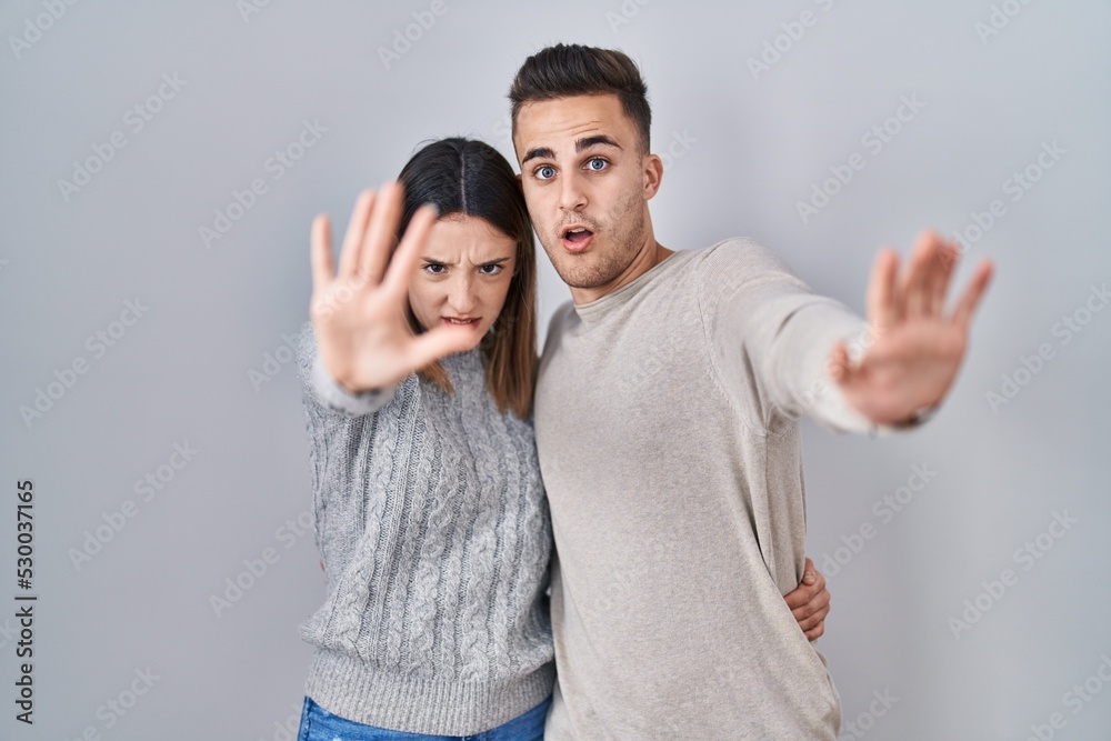 Young hispanic couple standing over white background doing stop gesture with hands palms, angry and frustration expression