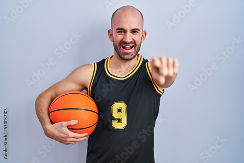 Young bald man with beard wearing basketball uniform holding ball pointing to you and the camera with fingers, smiling positive and cheerful