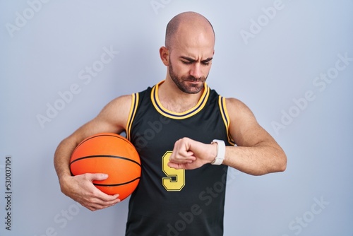 Young bald man with beard wearing basketball uniform holding ball checking the time on wrist watch, relaxed and confident