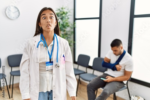 Young asian doctor woman at waiting room with a man with a broken arm making fish face with lips  crazy and comical gesture. funny expression.