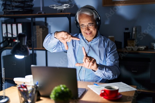 Hispanic senior man wearing call center agent headset at night smiling making frame with hands and fingers with happy face. creativity and photography concept.