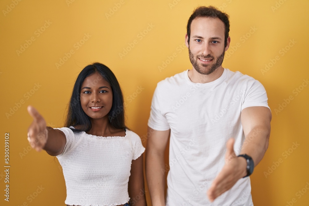 Interracial couple standing over yellow background smiling friendly offering handshake as greeting and welcoming. successful business. Interracial couple standing over yellow background smiling friendly offering handshake as greeting and welcoming. successful business.