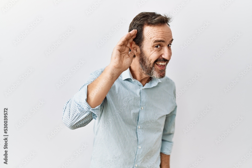 Middle age hispanic man with beard standing over isolated background smiling with hand over ear listening an hearing to rumor or gossip. deafness concept.