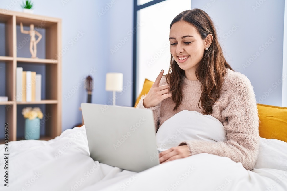 Young hispanic woman using computer laptop on the bed smiling happy pointing with hand and finger Young hispanic woman using computer laptop on the bed smiling happy pointing with hand and finger