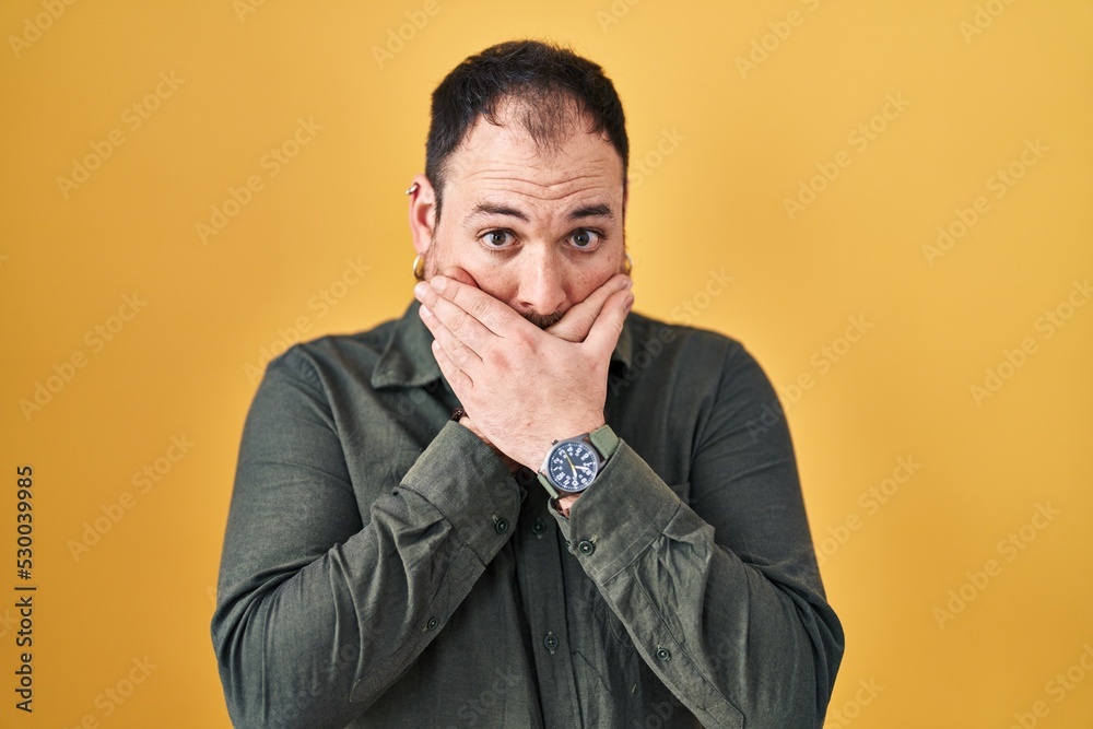 Plus size hispanic man with beard standing over yellow background shocked covering mouth with hands for mistake. secret concept. Plus size hispanic man with beard standing over yellow background shocked covering mouth with hands for mistake. secret concept.