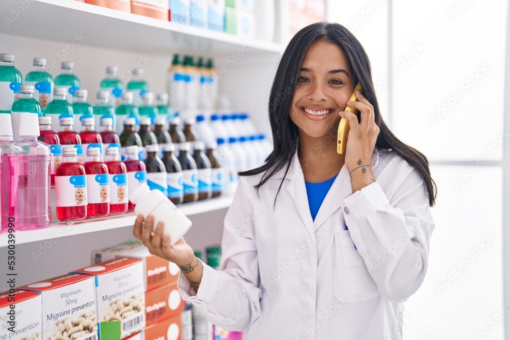 Young hispanic woman pharmacist holding pills bottle talking on smartphone at pharmacy