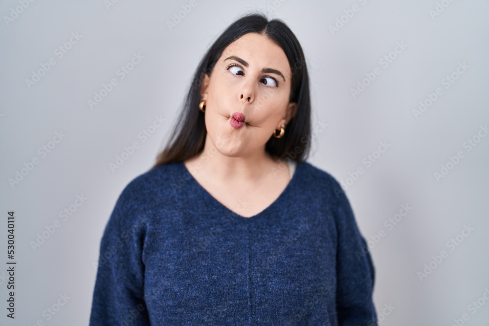 Obraz premium Young brunette woman standing over isolated background making fish face with lips, crazy and comical gesture. funny expression.