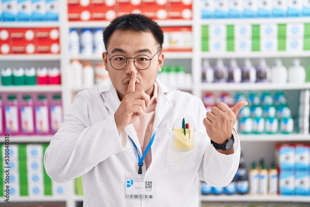 Chinese young man working at pharmacy drugstore asking to be quiet with finger on lips pointing with hand to the side. silence and secret concept. Chinese young man working at pharmacy drugstore asking to be quiet with finger on lips pointing with hand to the side. silence and secret concept.
