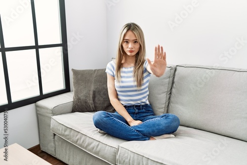 Asian young woman sitting on the sofa at home doing stop sing with palm of the hand. warning expression with negative and serious gesture on the face.