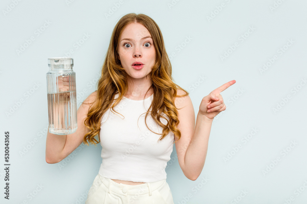 Young caucasian woman holding jar of water isolated on blue background pointing to the side Young caucasian woman holding jar of water isolated on blue background pointing to the side