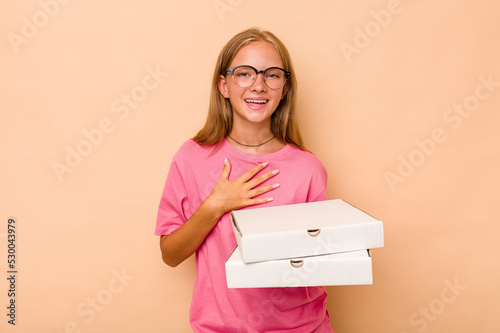 Little caucasian girl holding pizza isolated on beige background laughs out loudly keeping hand on chest.