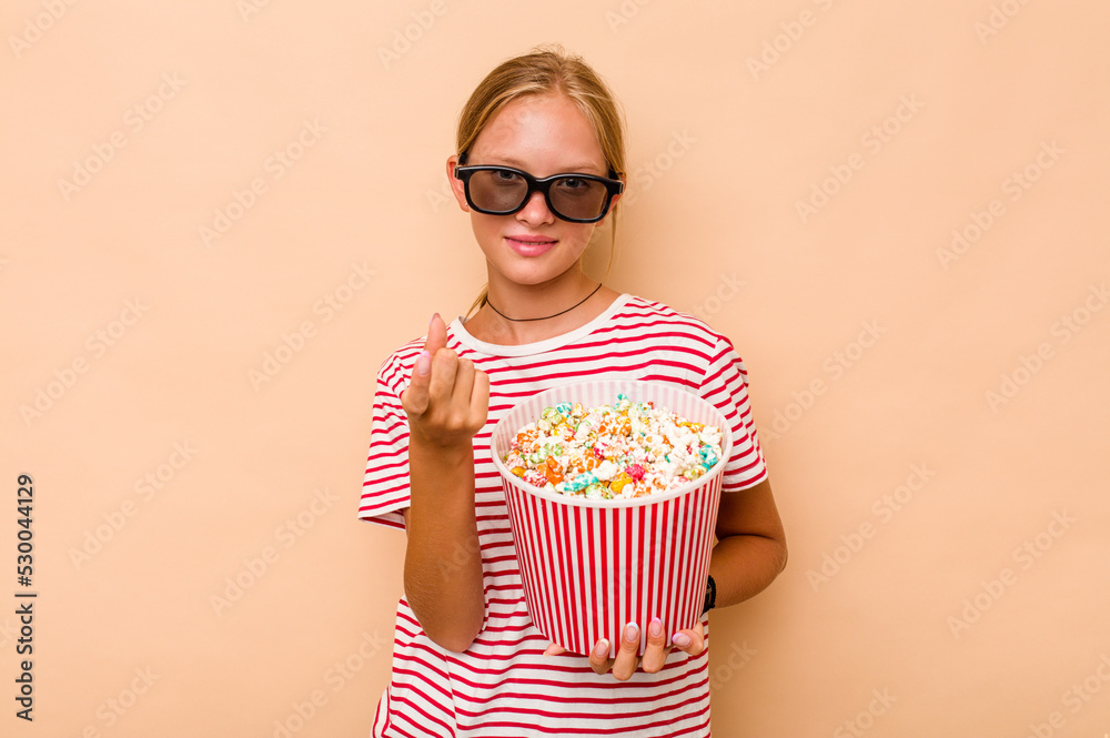 Obraz premium Little caucasian girl eating popcorn isolated on beige background pointing with finger at you as if inviting come closer.