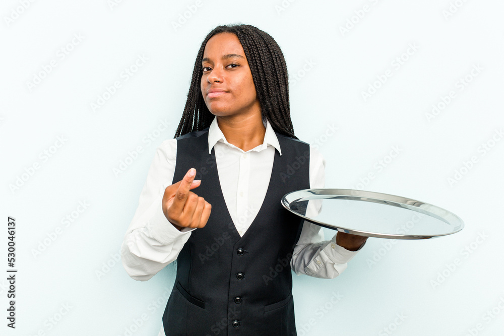 Young African American waitress woman holding a tray isolated on blue background pointing with finger at you as if inviting come closer. Young African American waitress woman holding a tray isolated on blue background pointing with finger at you as if inviting come closer.