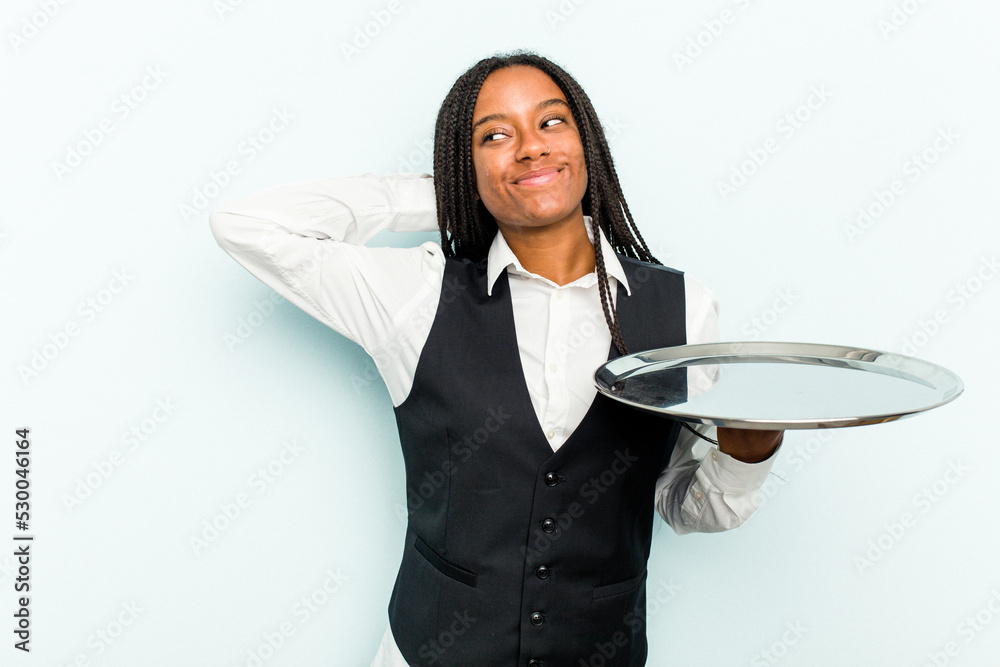 Young African American waitress woman holding a tray isolated on blue background touching back of head, thinking and making a choice. Young African American waitress woman holding a tray isolated on blue background touching back of head, thinking and making a choice.