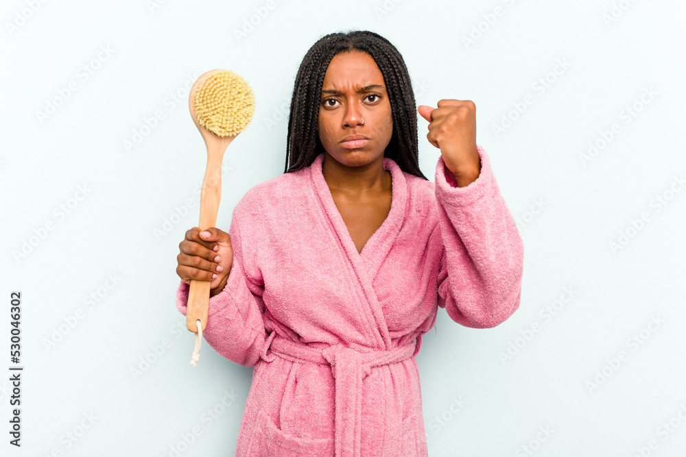 Young African American woman holding a bathtub brush isolated on blue background showing fist to camera, aggressive facial expression. Young African American woman holding a bathtub brush isolated on blue background showing fist to camera, aggressive facial expression.