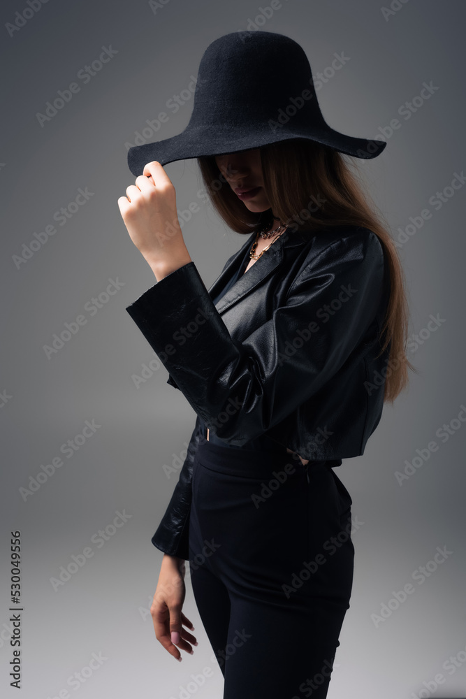brunette teenage model in black leather jacket adjusting floppy hat isolated on grey. brunette teenage model in black leather jacket adjusting floppy hat isolated on grey.