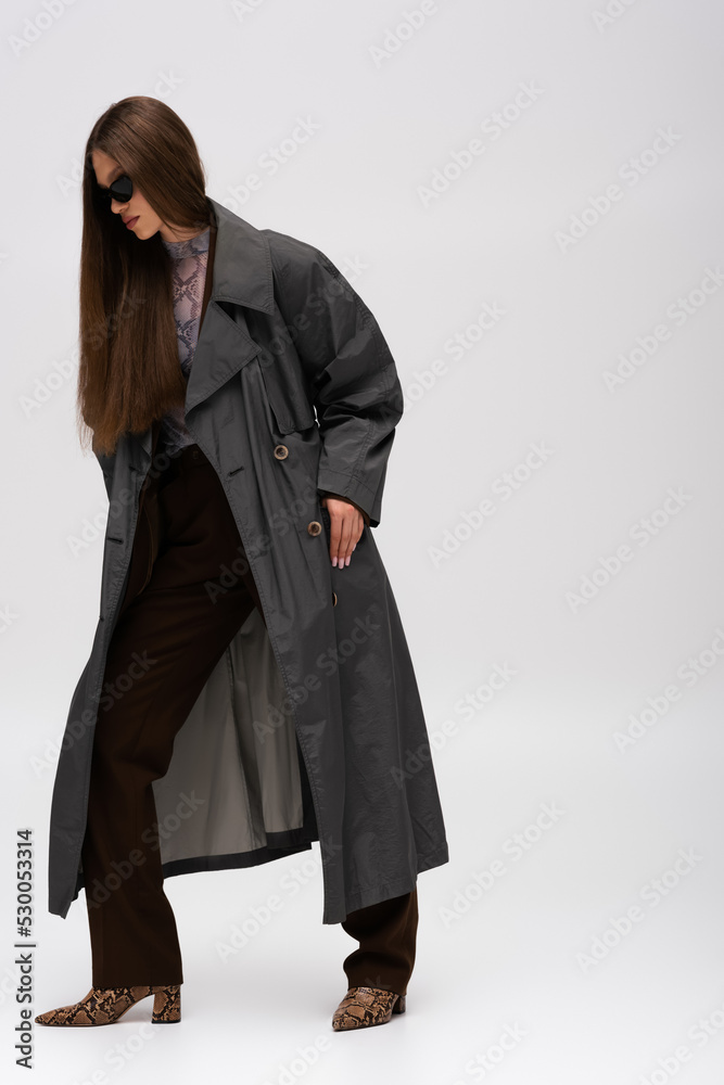 full length of teenage model in sunglasses and trench coat posing on gray. full length of teenage model in sunglasses and trench coat posing on gray.