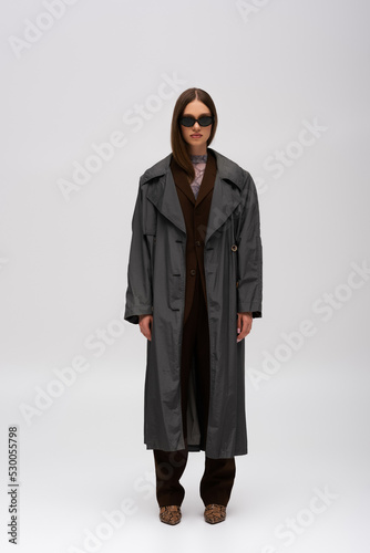 full length of model in stylish sunglasses and fashionable trench coat posing on grey.