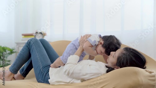 Healthy asian mother play with newborn baby in living room, mom holding lifting toddler on brown sofa with fun and tender. Adorable little boy having fun with mum togerther at home.
 photo