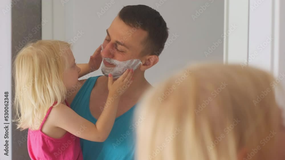 Little blonde girl joy and smears shaving foam on fathers face. Dad with daughter in arms smiling looking in mirror Little blonde girl joy and smears shaving foam on fathers face. Dad with daughter in arms smiling looking in mirror