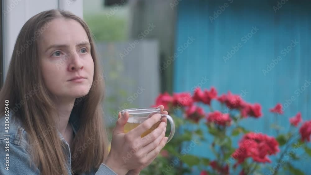 Tired young woman sitting outside house with tea, bush with flowers background. Brunette admires rainy weather Tired young woman sitting outside house with tea, bush with flowers background. Brunette admires rainy weather