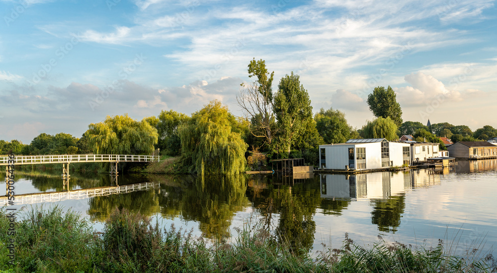 Obraz premium Village of Halfweg, municipality of Haarlemmermeer, The Netherlands, view of the wooden bridge and houseboats along the canal