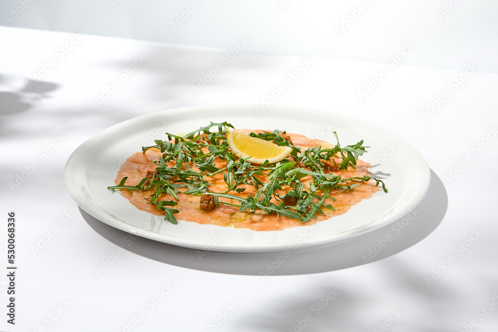 Salmon carpaccio with arugula on white background in summer day. Fish appetizer - salmon carpaccio in fine dining with shadows. Elegant carpaccio of raw salmon and sauce. Italian food. Aesthetic menu. Salmon carpaccio with arugula on white background in summer day. Fish appetizer - salmon carpaccio in fine dining with shadows. Elegant carpaccio of raw salmon and sauce. Italian food. Aesthetic menu.