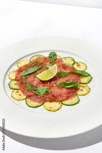 Beef carpaccio with zucchini and cucumber on white table with sunlight shadows. Appetizer of meat - beef carpaccio on summer dining. Sliced raw meat in italian style. Aesthetic menu.