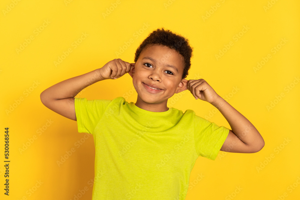 Portrait of funny preteen boy pulling his ears. Mixed race child wearing green T-shirt looking at camera and having fun against yellow background. Fun concept Portrait of funny preteen boy pulling his ears. Mixed race child wearing green T-shirt looking at camera and having fun against yellow background. Fun concept