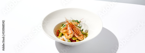 Elegant food - salad with shrimp and avocado on white table with harsh shadow. Flashy food concept. Seafood salad with shrimp, tomatoes and avocado on white plate with sunlight.