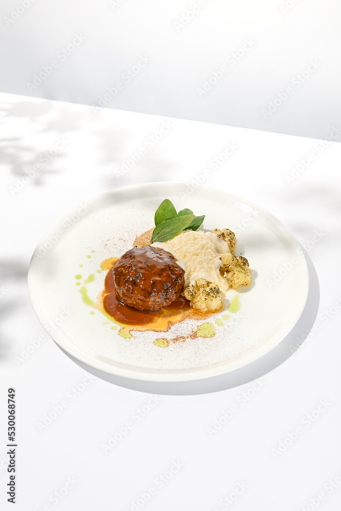 Beef cutlet with grilled cauliflower with gravy and cheese sauce on white plate. Elegant beefsteak on light background with shadows of sunlight. Summer menu with hard shadows Meat ball of minced meat Beef cutlet with grilled cauliflower with gravy and cheese sauce on white plate. Elegant beefsteak on light background with shadows of sunlight. Summer menu with hard shadows Meat ball of minced meat