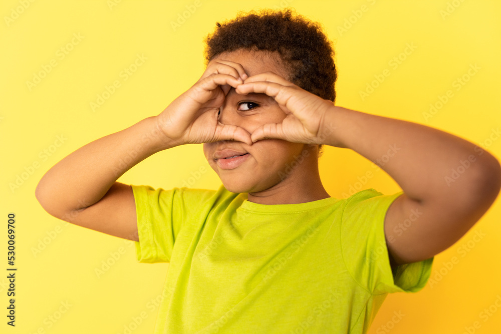 Portrait of happy preteen boy peeping through fingers. Mixed race child wearing green T-shirt looking at camera and smiling against yellow background. Fun concept Portrait of happy preteen boy peeping through fingers. Mixed race child wearing green T-shirt looking at camera and smiling against yellow background. Fun concept
