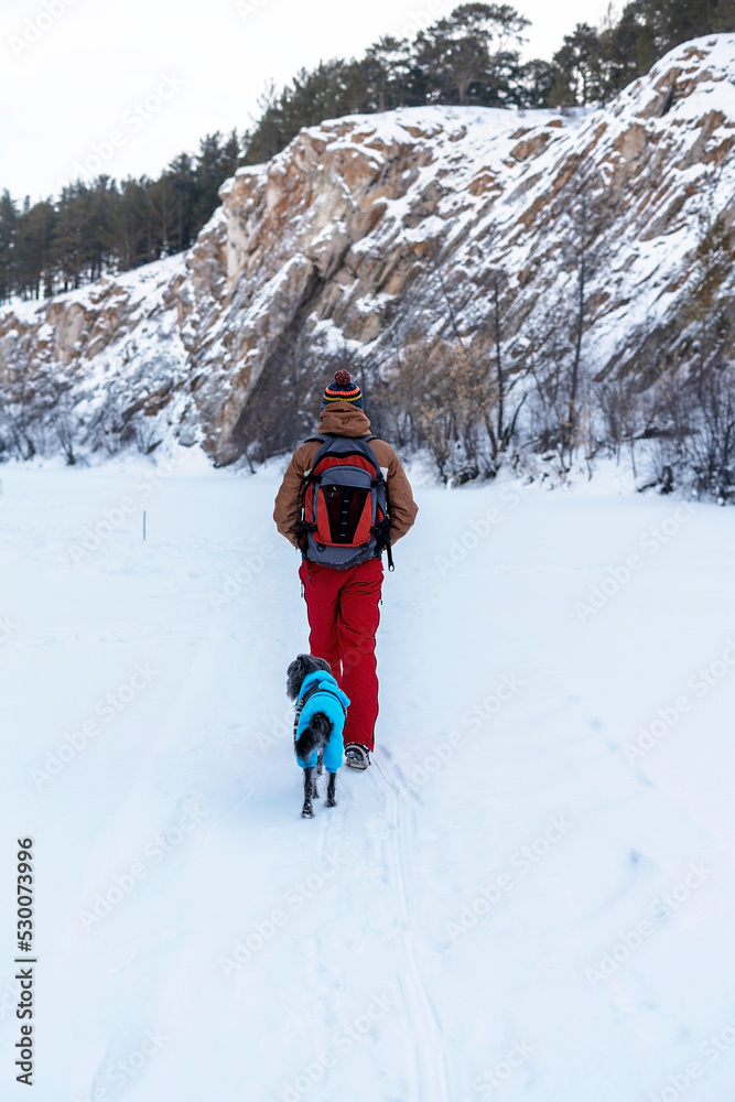 Rear view young man in red brown warm clothes with backpack with mixed breed dog in warm blue suit walking in snow among rocks and cliffs in winter Active lifestyle hiking pets adoption