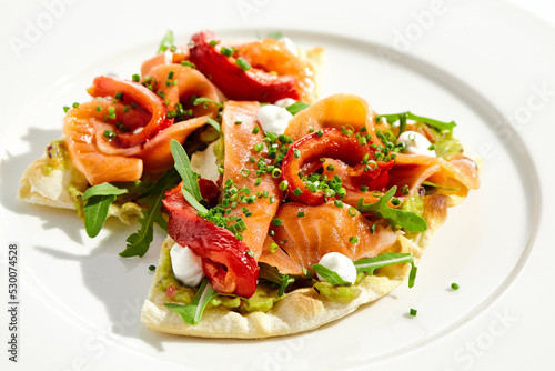 Bruschetta with salmon, avocado and paprika on white plate. Salmon bruschetta on crispy focaccia. Appetizer with guacamole, trout, cheese and paprika on toast. Fish antipasti in restaurant menu.