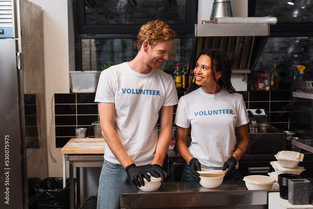 Obraz premium happy multiethnic volunteers in t-shirts preparing plastic containers with food in kitchen.