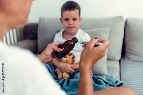 Schoolboy sick and home, willing to take some syrup from mom. Little boy taking a spoon of medicine.