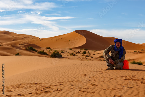 Fototapeta Naklejka Na Ścianę i Meble -  Man sitting on sand dunes and looking at the camera. Sahara Desert. Erg Chebbi dunes in Morocco